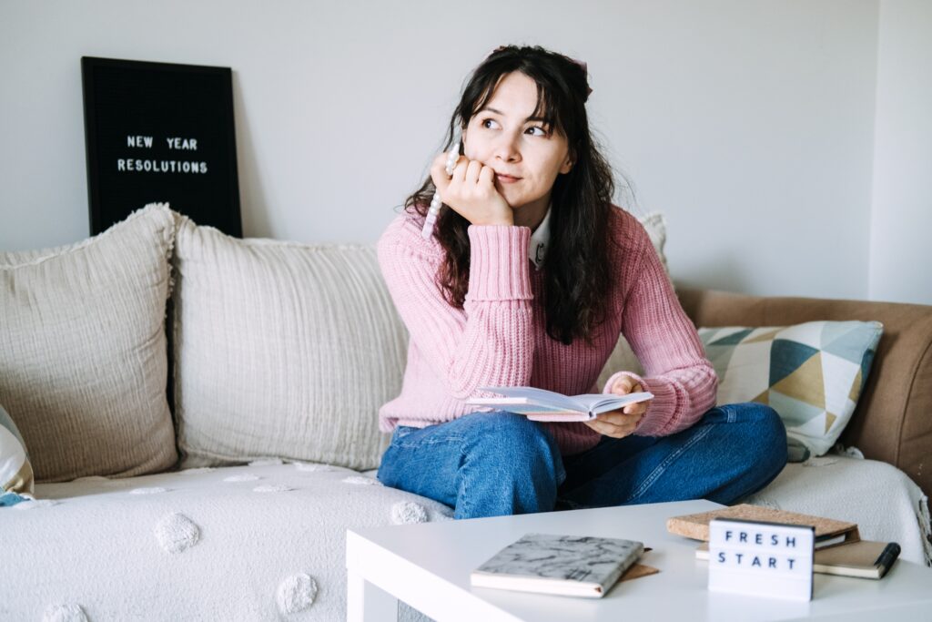 Young adult woman sits on sofa writing goals in small notebook