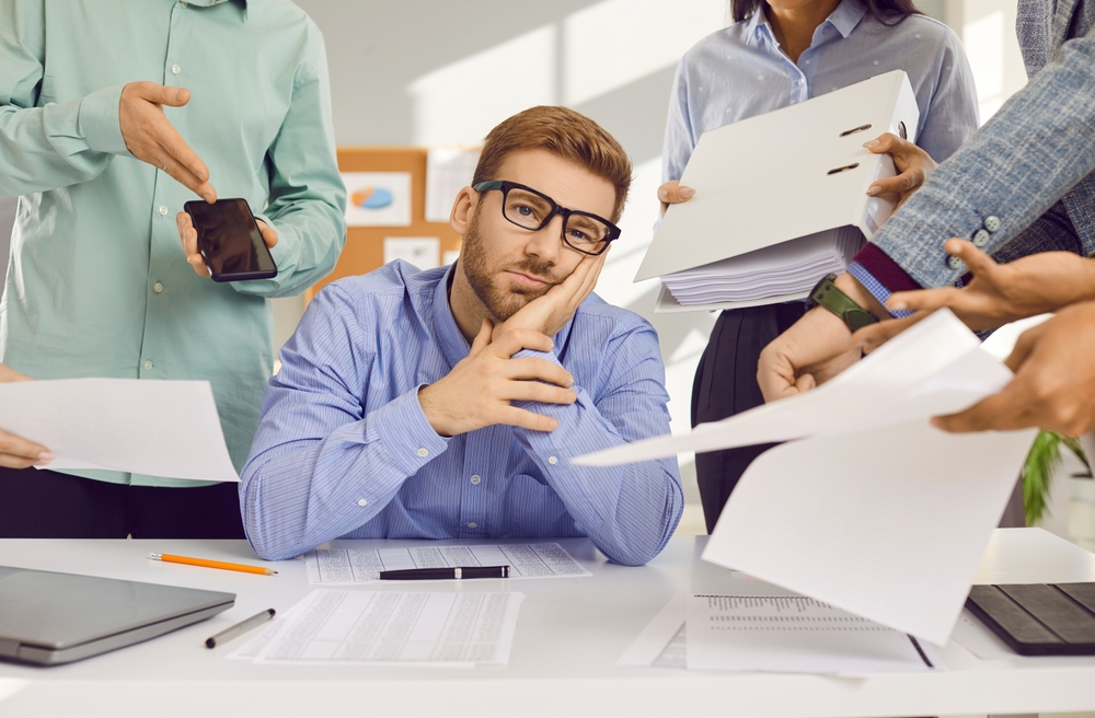 Stress, burnout and tired man sitting at the desk in office frustrated or overwhelmed by coworkers at workplace. Young businessman looking sad at camera. Overworked, multitasking concept.