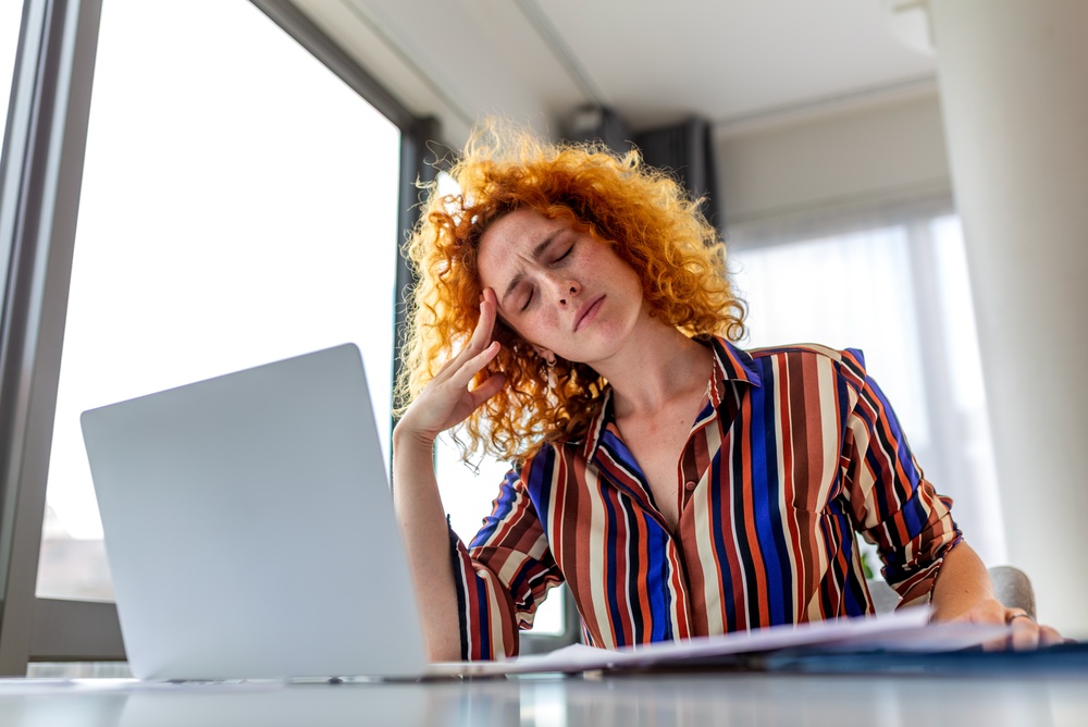 Exhausted redhead woman touching head with eyes closed suffering from migraine at home office desk.