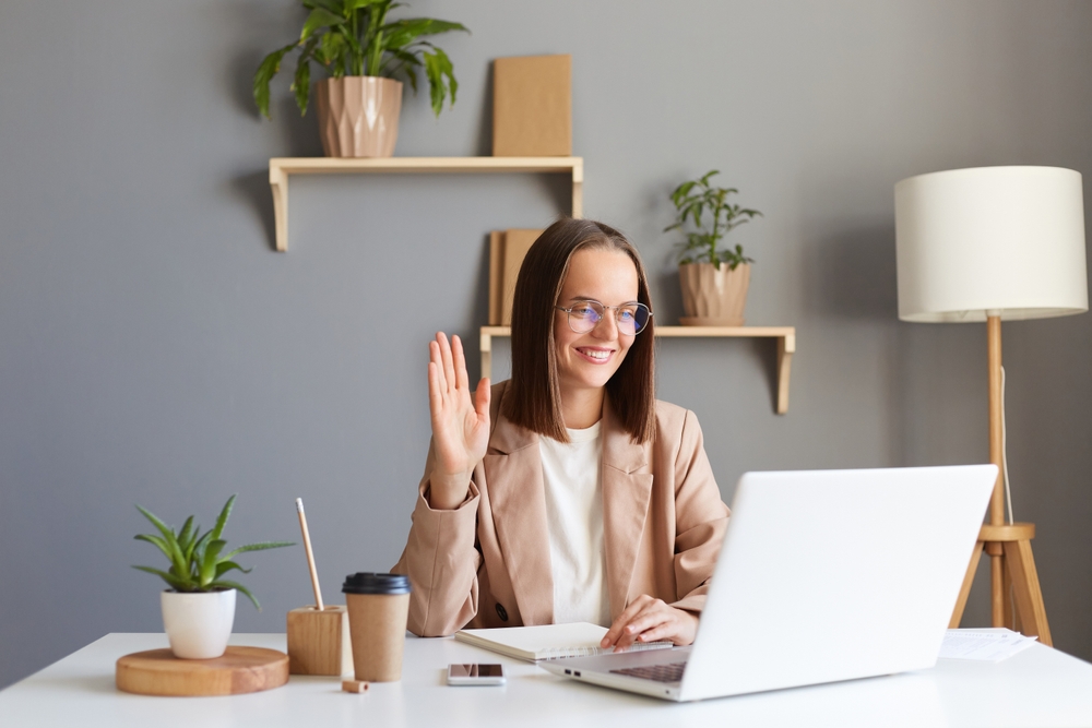 Indoor shot of business woman smiling friendly woman with brown hair wearing beige jacket working online on laptop, having video call, waving hand, saying hello or bye.